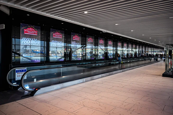 Sequenza di schermi promozionali lungo un corridoio aeroportuale con tapis roulant